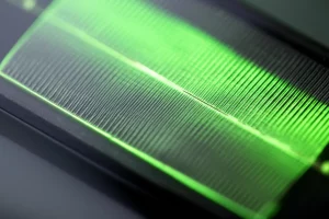 Primo piano fotorealistico di una cella solare organica flessibile e trasparente basata su nanofili d'argento, che mostra un bagliore verde tenue proveniente dallo strato di riciclo energetico plasmonico (PEEL). La cella è leggermente curvata per evidenziare la flessibilità. Obiettivo macro 90mm, illuminazione laterale controllata per evidenziare la texture sottile dei nanofili e la trasparenza generale, alta definizione, sfondo sfocato.