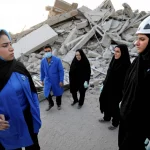 Fotografia realistica di un gruppo di infermieri iraniani, uomini e donne, che lavorano instancabilmente tra le rovine dopo il terremoto di Kermanshah. Stile reportage, luce naturale del tardo pomeriggio, obiettivo grandangolare 24mm per catturare l'ambiente devastato e lo sforzo collettivo, espressioni miste di fatica e determinazione.