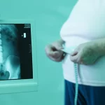 Fotografia medica concettuale: una scansione DXA della colonna vertebrale e del femore è visualizzata su uno schermo digitale, mentre in primo piano sfocato si vede una donna anziana che si misura il girovita con un metro a nastro. Lente prime 35mm, duotone blu e verde acqua per un'atmosfera clinica ma speranzosa, profondità di campo selettiva.