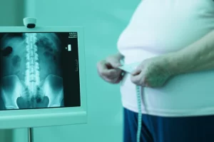 Fotografia medica concettuale: una scansione DXA della colonna vertebrale e del femore è visualizzata su uno schermo digitale, mentre in primo piano sfocato si vede una donna anziana che si misura il girovita con un metro a nastro. Lente prime 35mm, duotone blu e verde acqua per un'atmosfera clinica ma speranzosa, profondità di campo selettiva.