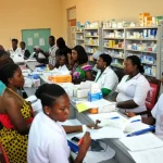 Fotografia realistica di una farmacia ambulatoriale affollata ma ordinata in un ospedale universitario nigeriano, obiettivo zoom 24-70mm a circa 35mm, luce ambientale mista, pazienti in attesa e farmacisti al lavoro dietro il bancone, focus nitido sui volti e sulle confezioni dei farmaci. Stile fotorealistico.