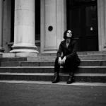 Ritratto fotografico in bianco e nero di una donna seduta sui gradini di un edificio urbano, sguardo perso nel vuoto, simboleggiando solitudine e resilienza. Obiettivo 35mm, profondità di campo ridotta per isolare il soggetto, stile film noir per accentuare il dramma.