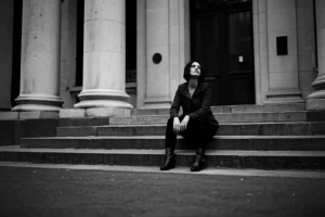 Ritratto fotografico in bianco e nero di una donna seduta sui gradini di un edificio urbano, sguardo perso nel vuoto, simboleggiando solitudine e resilienza. Obiettivo 35mm, profondità di campo ridotta per isolare il soggetto, stile film noir per accentuare il dramma.