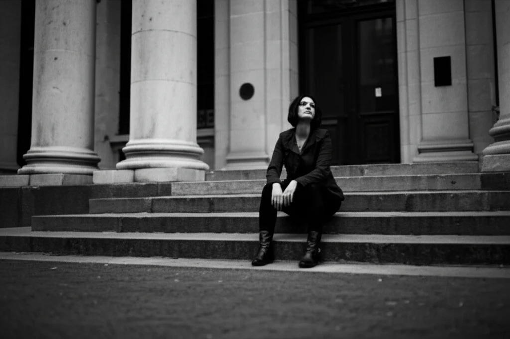 Ritratto fotografico in bianco e nero di una donna seduta sui gradini di un edificio urbano, sguardo perso nel vuoto, simboleggiando solitudine e resilienza. Obiettivo 35mm, profondità di campo ridotta per isolare il soggetto, stile film noir per accentuare il dramma.
