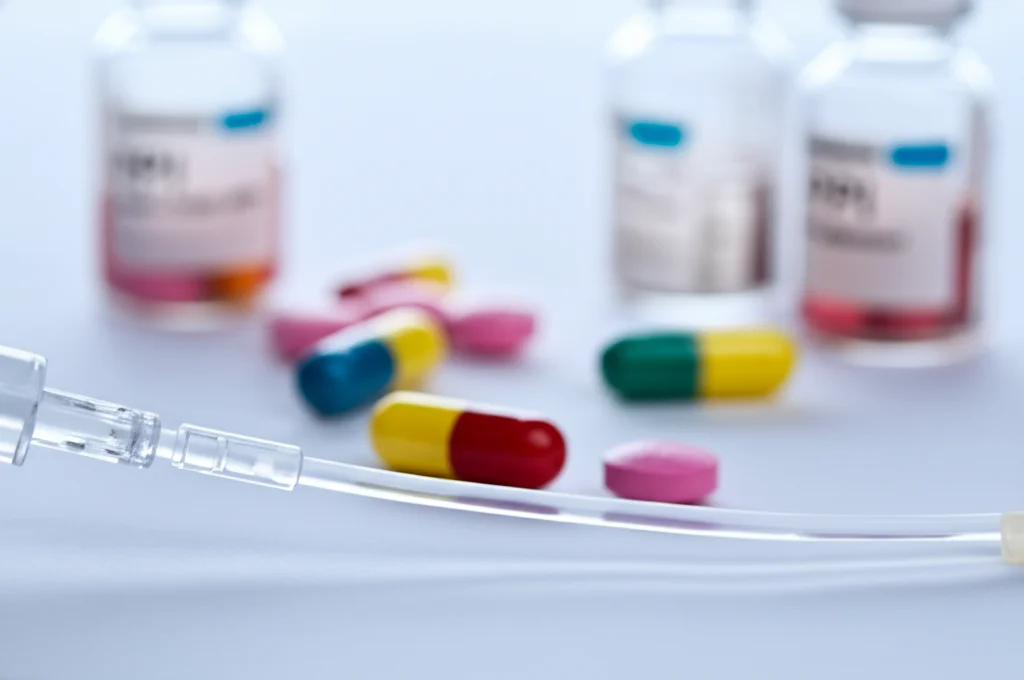 Fotografia macro (obiettivo 100mm) di una flebo per immunoterapia (ICI) in primo piano, con flaconi di pillole (PPI, antibiotici) sfocati sullo sfondo. Illuminazione controllata, alta definizione, focus preciso sulla flebo per simboleggiare l'interazione farmacologica nel trattamento del carcinoma uroteliale.
