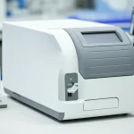Immagine fotorealistica del sistema diagnostico POCT iPonatic compatto su un banco di laboratorio pulito e moderno. La macchina è bianca e grigia, con una cartuccia diagnostica monouso inserita nello slot frontale e una luce LED verde accesa che indica il funzionamento. Accanto al dispositivo, un contagocce quantitativo iPonatic. Obiettivo prime 35mm, profondità di campo ridotta che sfoca leggermente lo sfondo del laboratorio (pipette, provette), mettendo a fuoco il dispositivo. Illuminazione chiara e professionale.