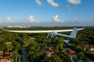 Immagine fotorealistica di un drone ad ala fissa bianco (simile al WingtraOne) che sorvola diagonalmente un quartiere residenziale ben curato di Coral Gables, con case unifamiliari dai tetti rossi e strade alberate. La luce è quella calda del tardo pomeriggio, che crea ombre lunghe e definite. Obiettivo zoom teleobiettivo 100mm per comprimere leggermente la prospettiva e dare un senso di osservazione dettagliata, messa a fuoco nitida sul drone e sul quartiere sottostante, cielo azzurro con qualche nuvola.