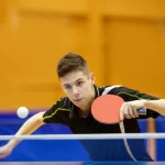 Atleta di tennistavolo in piena azione durante un colpo di dritto, focus sulla coordinazione braccio-racchetta e sull'espressione concentrata. Teleobiettivo 200mm, fast shutter speed per catturare il movimento rapido della pallina e della racchetta, sfondo della palestra leggermente sfocato per enfatizzare il giocatore.