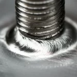 Fotografia macro realistica, obiettivo 100mm, di un dettaglio ravvicinato della testa di un bullone in acciaio saldato su una lamiera metallica. Messa a fuoco sulla texture irregolare del cordone di saldatura ad arco che collega la base del bullone alla lamiera. Alta definizione, messa a fuoco precisa, illuminazione controllata che enfatizza le superfici metalliche e le imperfezioni della saldatura. Contesto industriale suggerito.