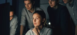 Fotografia stile film noir, 35mm, di un gruppo diversificato di sviluppatori software che collaborano attorno a uno schermo luminoso in un ufficio moderno poco illuminato, profondità di campo ridotta focalizzata sul volto concentrato di una persona che sembra guidare la discussione, toni blu e grigi duotone, simboleggiando la leadership e la dinamica di squadra nello sviluppo software.