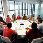 Una sala conferenze moderna e luminosa, vista grandangolare, con diverse persone di etnie e età varie che discutono animatamente attorno a un tavolo rotondo pieno di appunti, simboleggiando la collaborazione scientifica e lo scambio di idee promossi da McMahon. Obiettivo 24mm, luce naturale diffusa dalle finestre.