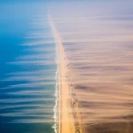 Vista aerea grandangolare, 18mm, dell'Africa Occidentale che mostra le vaste distese del deserto del Sahara che sfumano verso la savana e la costa atlantica, con visibili pennacchi di polvere desertica trasportati dai venti verso l'oceano, lunga esposizione per suggerire il movimento atmosferico, colori naturali vividi e messa a fuoco nitida sull'intera scena.