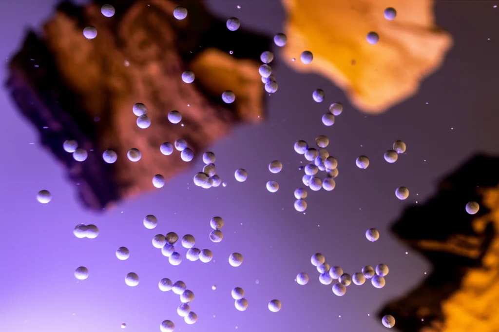 Immagine fotorealistica che mostra nanoparticelle sferiche di ZnO sospese in acqua limpida sotto una luce UV stilizzata, con frammenti di corteccia di Cuachalalate visibili sullo sfondo sfocato. Obiettivo macro 80mm, alta definizione, illuminazione drammatica che evidenzia le particelle e l'effetto fotocatalitico, profondità di campo ridotta.