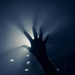 Immagine simbolica potente: una mano tesa appartenente a un adolescente di etnia Nera o Latina che emerge dall'ombra scura verso una luce fioca e speranzosa, rappresentando la difficile ricerca di aiuto per la salute mentale. Stile fotografico realistico e drammatico, 35mm lens, contrasto elevato tra luci e ombre, duotone blu scuro e grigio chiaro per enfatizzare l'emozione, profondità di campo che sfoca lo sfondo per concentrarsi sulla mano e sulla luce.