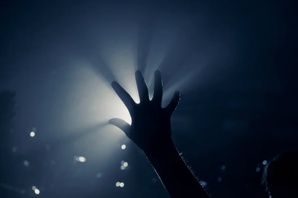 Immagine simbolica potente: una mano tesa appartenente a un adolescente di etnia Nera o Latina che emerge dall'ombra scura verso una luce fioca e speranzosa, rappresentando la difficile ricerca di aiuto per la salute mentale. Stile fotografico realistico e drammatico, 35mm lens, contrasto elevato tra luci e ombre, duotone blu scuro e grigio chiaro per enfatizzare l'emozione, profondità di campo che sfoca lo sfondo per concentrarsi sulla mano e sulla luce.