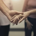 Fotografia simbolica, due mani diverse che si stringono delicatamente, una offre supporto all'altra, sfondo leggermente sfocato di un ambiente comunitario o clinico accogliente, obiettivo 50mm, profondità di campo ridotta, luce calda e morbida, toni duo seppia e grigio.