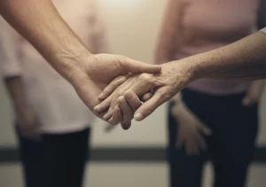 Fotografia simbolica, due mani diverse che si stringono delicatamente, una offre supporto all'altra, sfondo leggermente sfocato di un ambiente comunitario o clinico accogliente, obiettivo 50mm, profondità di campo ridotta, luce calda e morbida, toni duo seppia e grigio.