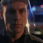 Immagine fotorealistica di un paramedico in primo piano, visiera protettiva alzata, gocce di sudore sulla fronte, sguardo intenso e focalizzato oltre l'obiettivo, sullo sfondo sfocato le luci di un'emergenza. Obiettivo prime 35mm, profondità di campo, leggero effetto film noir.