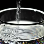 Primo piano fotorealistico di acqua che scorre da un rubinetto metallico in un bicchiere di vetro trasparente. Nell'acqua sono visibili minuscoli puntini colorati che rappresentano microplastiche e una leggera iridescenza che suggerisce la presenza di metalli disciolti. Lente macro 100mm, profondità di campo ridotta, illuminazione laterale drammatica, alta definizione.