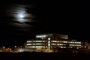 Immagine notturna suggestiva della luna piena che splende sopra il profilo di un pronto soccorso ospedaliero moderno. Fotografia astronomica, obiettivo grandangolare 15mm, lunga esposizione per enfatizzare la luce lunare, focus nitido.
