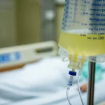 Primo piano di una sacca di drenaggio ascitico parzialmente riempita accanto a un letto d'ospedale, luce clinica controllata, obiettivo macro 60mm, alta definizione, messa a fuoco sul liquido giallastro, simbolo delle cure palliative per l'ascite refrattaria.