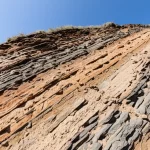 Sezione trasversale geologica realistica che mostra strati rocciosi inclinati con un evidente strato intermedio debolmente cementato. Wide-angle lens, 15mm, sharp focus, natural lighting highlighting rock texture.