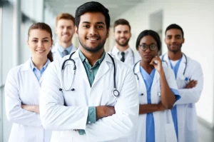 Fotografia di un gruppo diversificato di studenti di medicina sorridenti ma dall'aria stanca, in camice bianco, in piedi in un corridoio luminoso di un ospedale universitario. Obiettivo zoom 24-70mm a circa 35mm, luce naturale morbida, profondità di campo media per mostrare sia i volti che l'ambiente.