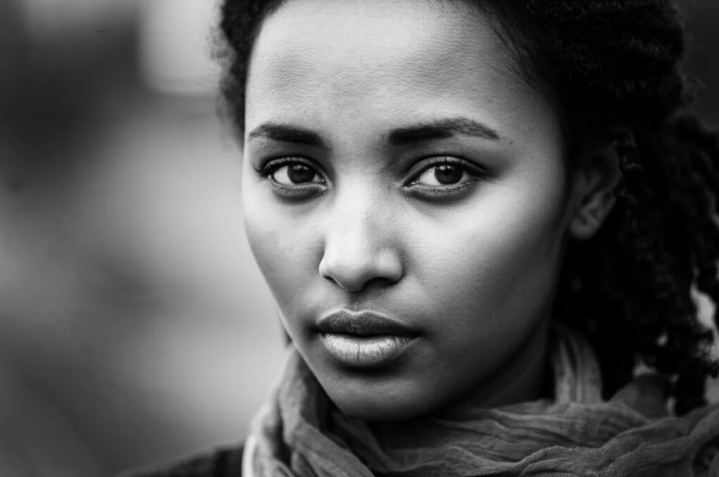 Ritratto fotografico intenso di una giovane donna etiope, sguardo fiducioso e diretto verso l'obiettivo, luce laterale morbida che ne illumina metà volto creando contrasto, obiettivo 50mm, profondità di campo ridotta che sfoca lo sfondo, bianco e nero stile film noir per enfatizzare l'espressione determinata.