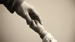 Immagine fotorealistica che mostra una mano adulta che tiene delicatamente una piccola mano di bambino, in bianco e nero con toni seppia, obiettivo 35mm con profondità di campo, simboleggiando la vulnerabilità e la speranza nel trattamento di malattie pediatriche gravi come la SMA.