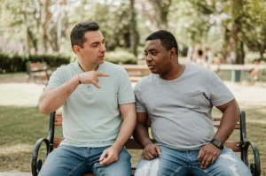 Due uomini, uno di etnia caucasica e uno afroamericano, entrambi sulla quarantina e visibilmente sovrappeso, seduti fianco a fianco su una panchina in un parco cittadino, in una conversazione seria ma non tesa. Luce naturale morbida. Obiettivo prime 35mm, profondità di campo media per mantenere a fuoco entrambi i soggetti ma sfocare leggermente lo sfondo. Stile fotorealistico.