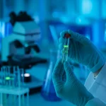 Primo piano fotorealistico, obiettivo prime 35mm, che mostra le mani guantate di uno scienziato/a che tengono con cura una fiala contenente una soluzione di nanoparticelle luminescenti (leggero bagliore verde-bluastro che rappresenta l'ICG). Attrezzature di laboratorio (microscopio, provette) sono sfocate sullo sfondo. Profondità di campo ridotta. L'immagine suggerisce ricerca avanzata sul cancro, precisione e speranza.