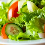 Un primo piano invitante di un'insalata mista fresca e colorata in una ciotola bianca, fotografata con obiettivo macro 100mm per alta definizione dei dettagli delle verdure (lattuga, pomodoro, cetriolo). L'illuminazione è controllata ma proietta un'ombra sottile sul bordo del piatto, suggerendo un pericolo nascosto o una contaminazione non visibile, simboleggiando i risultati dello studio sulla contaminazione microbica in Ghana.