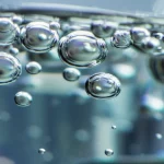 Macro fotografia (obiettivo macro 100mm) che cattura nanobolle di CO2 iridescenti mentre salgono attraverso acqua limpida all'interno di un reattore ad alta pressione in acciaio, alto dettaglio, messa a fuoco precisa, illuminazione scientifica controllata che evidenzia le bolle.