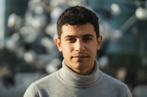 Primo piano ritratto fotografico di una persona che guarda con sicurezza davanti a sé, leggermente sorridente, con uno sfondo sfocato che suggerisce connessioni sociali; 50mm prime lens, depth of field, luce naturale calda.
