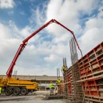 Pompa per calcestruzzo moderna in funzione in un grande cantiere edile, getto di calcestruzzo visibile che esce dal braccio articolato verso una cassaforma in altezza, operai con caschi e giubbotti ad alta visibilità nelle vicinanze, luce del giorno intensa e cielo parzialmente nuvoloso, grandangolo 24mm, messa a fuoco nitida sull'azione, leggero motion blur sul calcestruzzo in movimento.