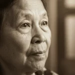 Ritratto fotografico di una persona anziana di origine asiatica che guarda con speranza verso una fonte di luce, simboleggiando il successo del trattamento del glaucoma (trabeculo-canalectomia) e il recupero della vista. Lente prime 35mm, profondità di campo ridotta per focalizzare sugli occhi espressivi, toni caldi e luminosi, bianco e nero con viraggio seppia leggero.