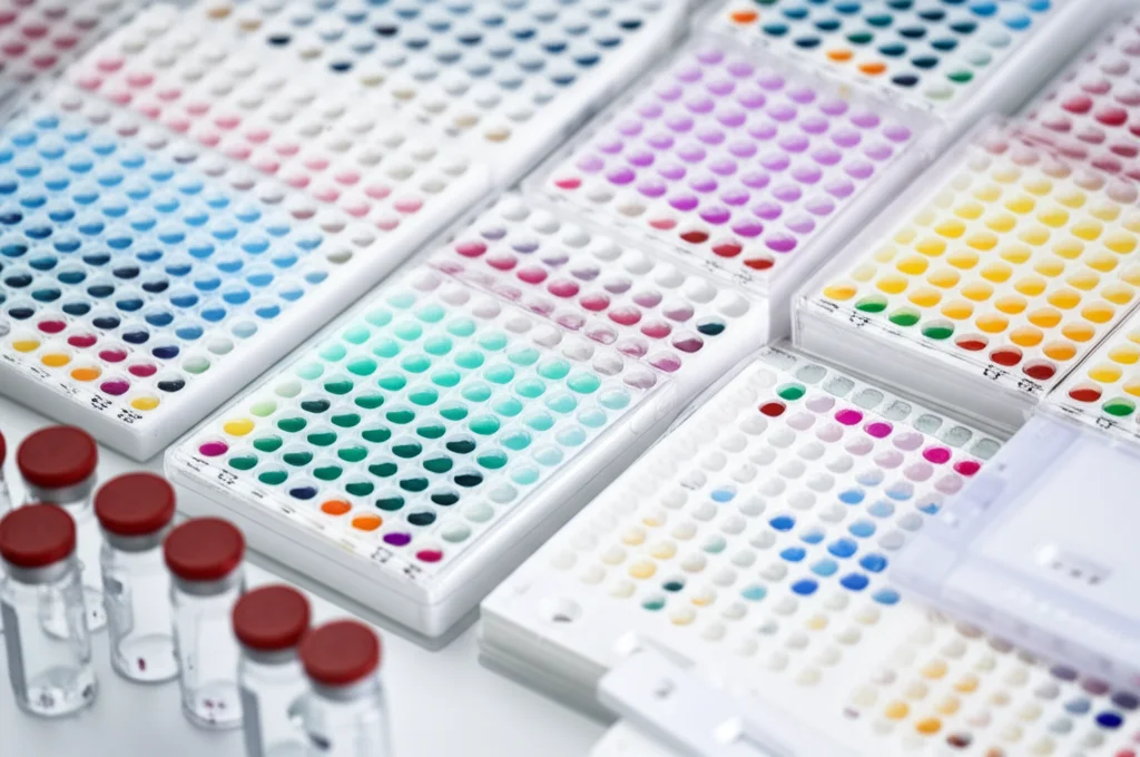 Fotografia macro, obiettivo 90mm, di piastre microtiter ELISA con pozzetti colorati che indicano risultati, affiancate a cassette di test rapidi e fiale, suggerendo un confronto tra test anticorpali SARS-CoV-2. Illuminazione controllata, alto dettaglio.