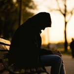 Immagine concettuale fotorealistica: primo piano sfocato di una persona seduta da sola su una panchina in un parco urbano al tramonto, con lo sguardo perso nel vuoto, simboleggiando l'isolamento e il trauma psicologico post-violenza armata. Obiettivo 50mm, profondità di campo ridotta, luce naturale calda e filtrata dagli alberi, toni malinconici, alta definizione.