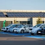 Foto panoramica realistica di un parcheggio aziendale moderno con auto elettriche collegate a colonnine di ricarica intelligenti, pannelli solari visibili sul tetto dell'edificio in background. Obiettivo grandangolare 24mm, luce del tardo pomeriggio, messa a fuoco nitida su tutto il panorama.