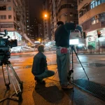Scena notturna cinematografica nelle strade trafficate e illuminate al neon di Hong Kong, asfalto bagnato che riflette le luci, una piccola troupe cinematografica indipendente al lavoro con attrezzature visibili, stile film noir, obiettivo grandangolare 20mm, lunga esposizione per catturare il movimento delle luci, messa a fuoco nitida sulla troupe.