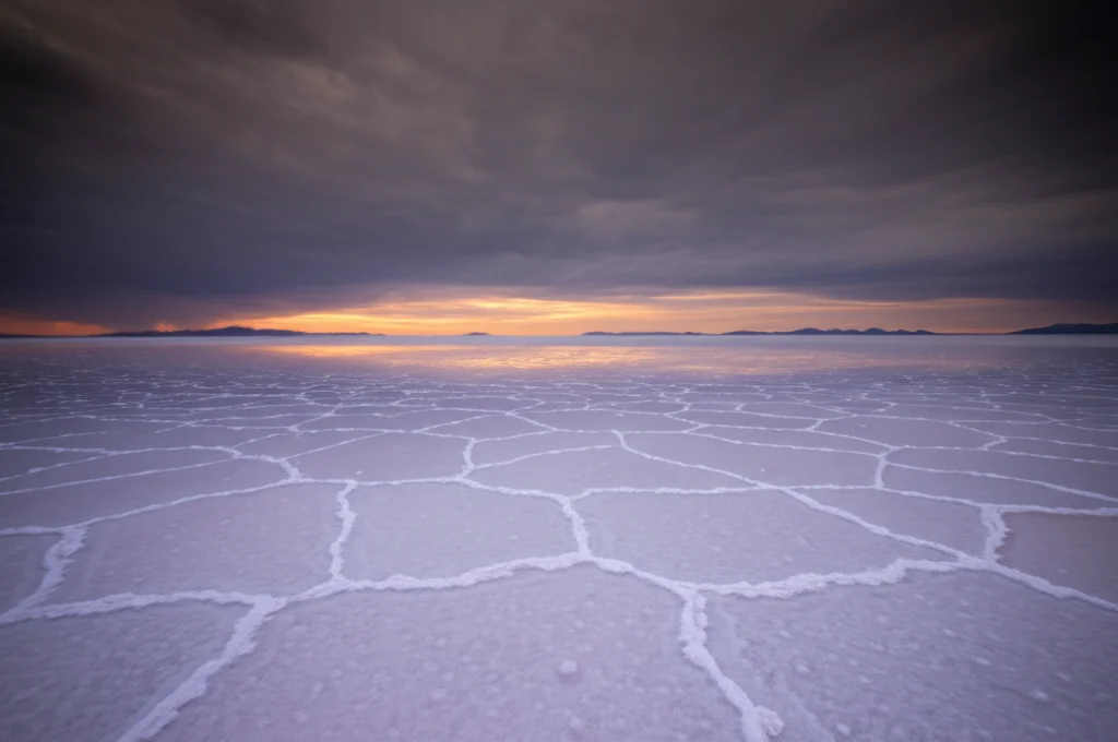 Paesaggio panoramico del Lago Salato di Qarhan, Cina. Vista grandangolare (10mm) che cattura l'immensità delle saline bianche che si estendono fino all'orizzonte, sotto un cielo drammatico al tramonto o all'alba. Focus nitido su tutta la scena, lunga esposizione per creare un effetto liscio sull'acqua residua e sulle nuvole. Colori caldi del cielo contrastano con il bianco del sale.