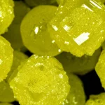 Fotografia macro (obiettivo 100mm) di brillanti cristalli gialli di YLID, alcuni sferici e altri di forma naturale, disposti su una superficie scura. Illuminazione controllata che evidenzia le sfaccettature e la struttura cristallina, alta definizione dei dettagli.