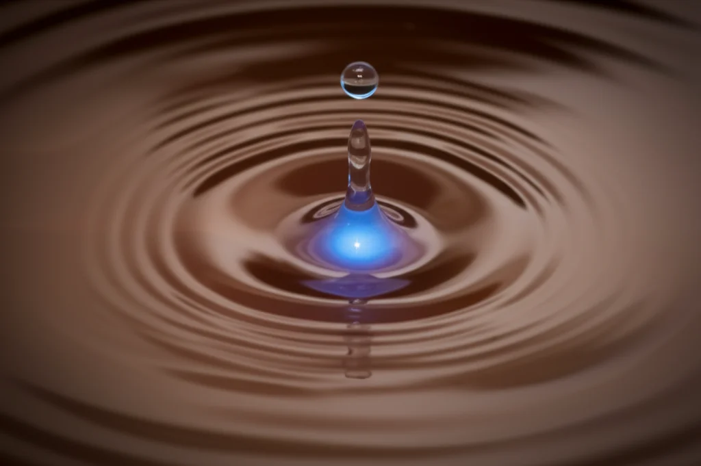 Immagine fotorealistica di una goccia d'acqua limpida che cade in acqua leggermente torbida, creando increspature; al centro della goccia, una debole luminescenza blu simboleggia l'azione enzimatica della laccasi. Obiettivo macro 90mm, illuminazione drammatica controllata, alta definizione.