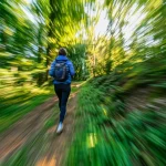 Una persona cammina a passo svelto su un sentiero immerso nel verde durante una giornata di sole, vista da dietro, obiettivo zoom 50mm, effetto leggermente mosso per indicare movimento, colori vivaci della natura, profondità di campo media che lascia intravedere il paesaggio.