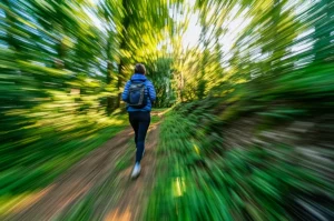 Una persona cammina a passo svelto su un sentiero immerso nel verde durante una giornata di sole, vista da dietro, obiettivo zoom 50mm, effetto leggermente mosso per indicare movimento, colori vivaci della natura, profondità di campo media che lascia intravedere il paesaggio.