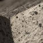 Fotografia still life, obiettivo macro 80mm, di un campione di calcestruzzo geopolimerico ad alta resistenza rinforzato con fibre di basalto e polvere di pomice vulcanica, illuminazione controllata che ne esalta la texture compatta e le fibre integrate, alta definizione, messa a fuoco precisa sul materiale innovativo.