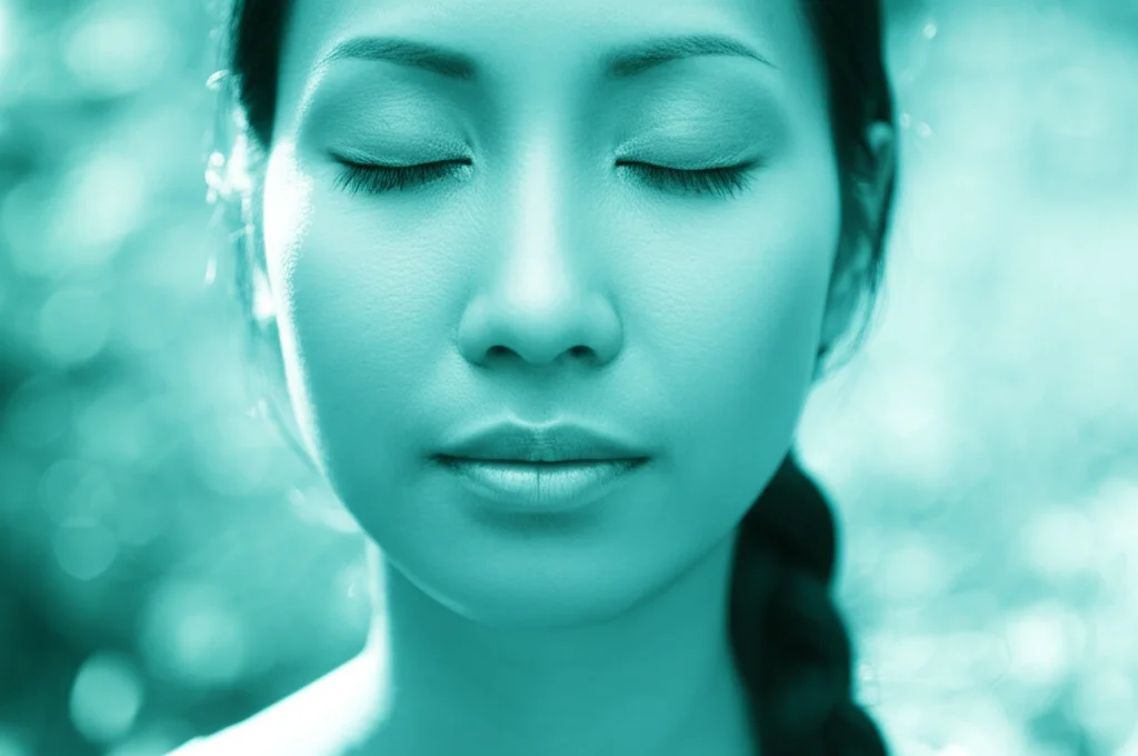 Immagine evocativa del benessere mentale e della tranquillità interiore. Fotografia ritratto, 35mm, una persona con gli occhi chiusi e un'espressione serena, immersa in una luce naturale morbida. Toni duotone blu e verde acqua, profondità di campo che sfoca delicatamente lo sfondo naturale, simboleggiando l'equilibrio raggiunto tramite la mindfulness e la tranquillità.