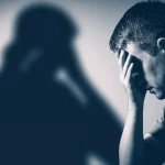 Immagine simbolica che rappresenta il legame tra un passato difficile (infanzia, magari rappresentato da un'ombra o un elemento sfocato) e l'ansia presente (un adulto con espressione tesa o preoccupata), con un elemento di speranza o resilienza (un raggio di luce, una pianta che cresce). Fotografia 35mm, toni duotone blu e grigio, profondità di campo.