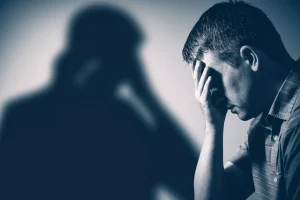 Immagine simbolica che rappresenta il legame tra un passato difficile (infanzia, magari rappresentato da un'ombra o un elemento sfocato) e l'ansia presente (un adulto con espressione tesa o preoccupata), con un elemento di speranza o resilienza (un raggio di luce, una pianta che cresce). Fotografia 35mm, toni duotone blu e grigio, profondità di campo.