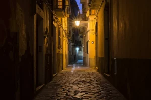 Un vicolo siciliano al tramonto, obiettivo grandangolare 20mm, luce lunga e ombre profonde che si estendono sulla pavimentazione antica, evocando il controllo territoriale persistente ma sfuggente della mafia attraverso l'evoluzione del pizzo.